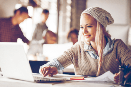 A young woman sitting alone at a worktable in an office reviewing hard copy designs and comparing them with content on a laptop,