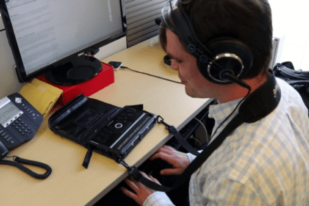 Man at a desk wearing headphones sitting before a computer using a braille keyboard.