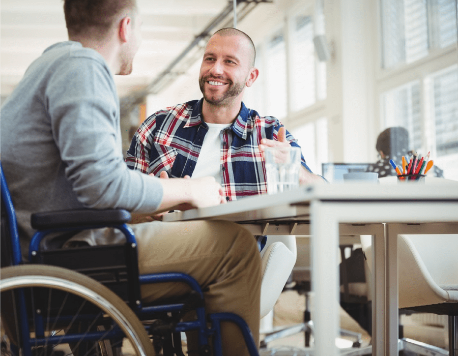 Two men in an office having an informal conversation around a desk. One of the men is in a wheelchair.