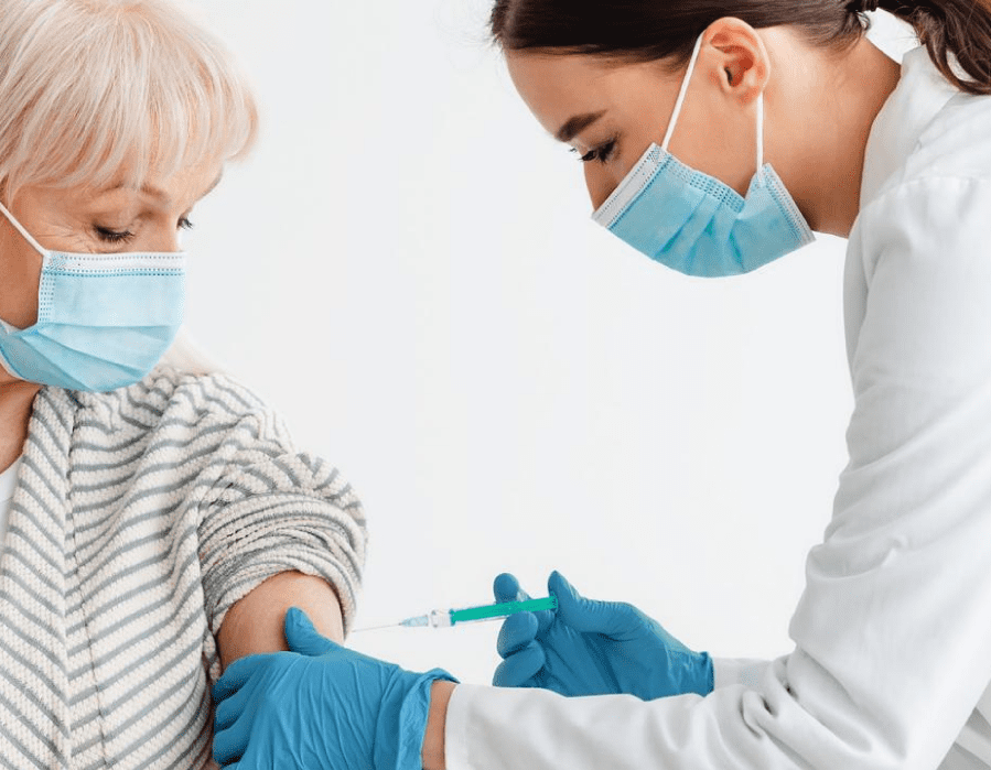 A female doctor giving an elderly woman a shot in her arm. Both doctor and patient are wearing surgical-style masks.