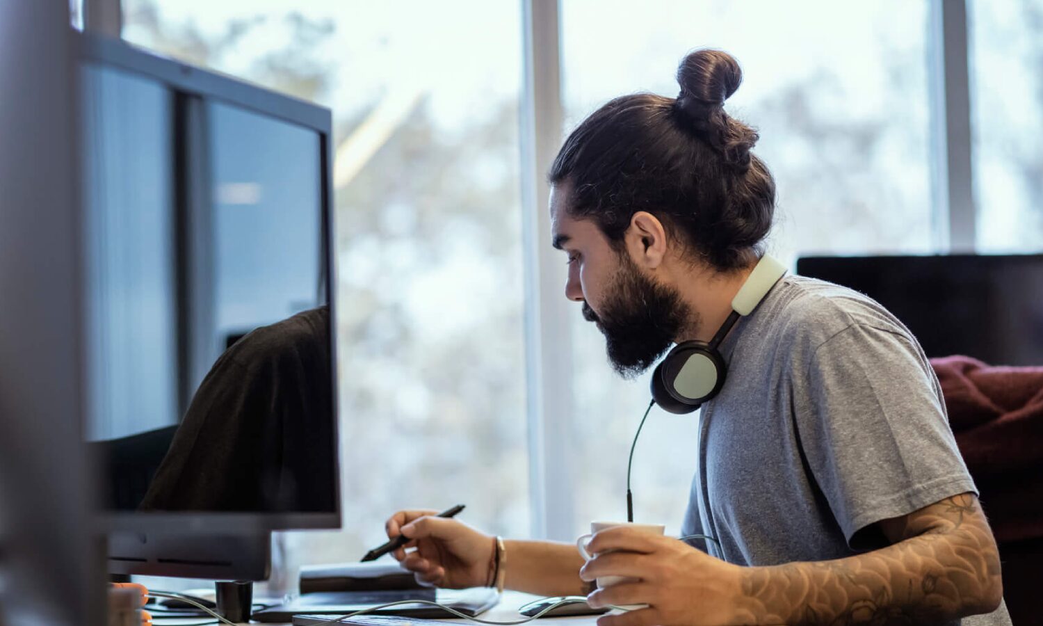 Man at his desk reviewing documents while working on his computer. He has a cup of coffee in his left hand.