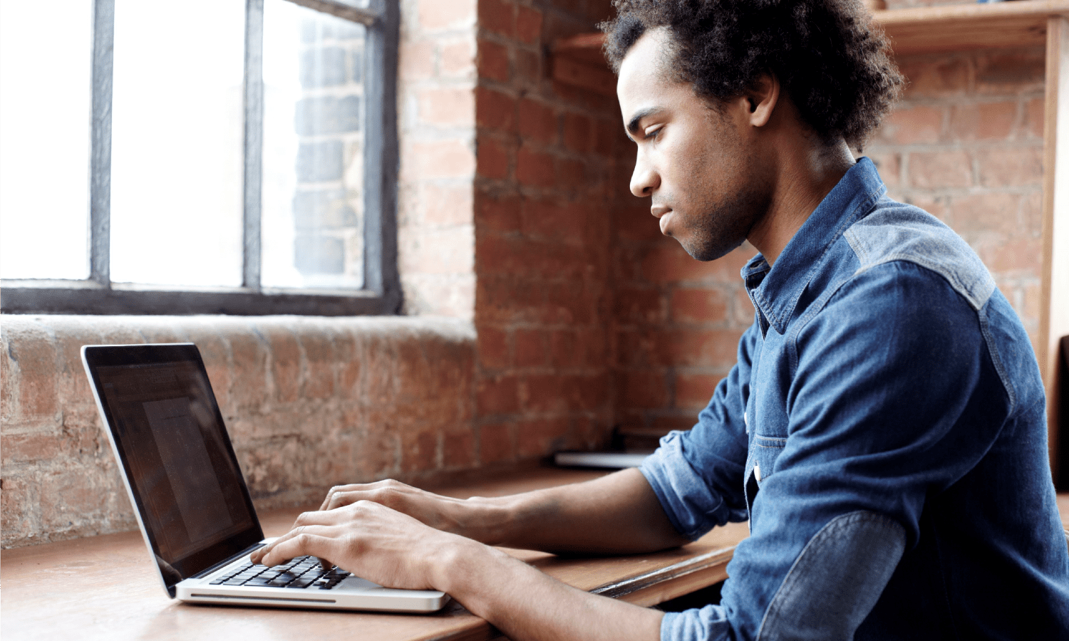 Young African Americal man working on a laptop at a desk near a window.