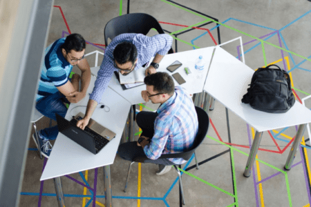 Three co-workers gathered around geometric tables in an open workspace looking at content on a laptop.