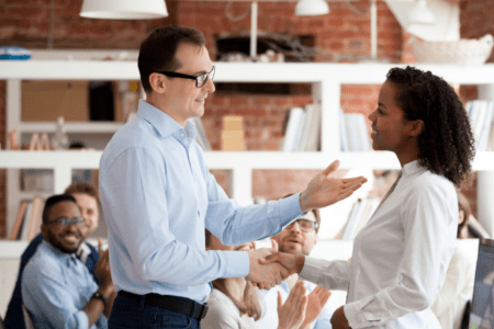 Two employees shaking hands while seated co-workers applaud.