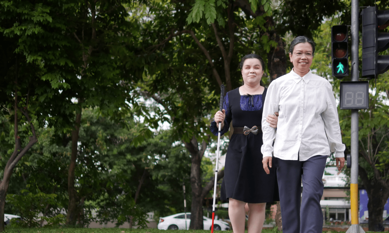 Two women crossing a street at a crosswalk. One of the women is helping the other who is using a blind guide cane.