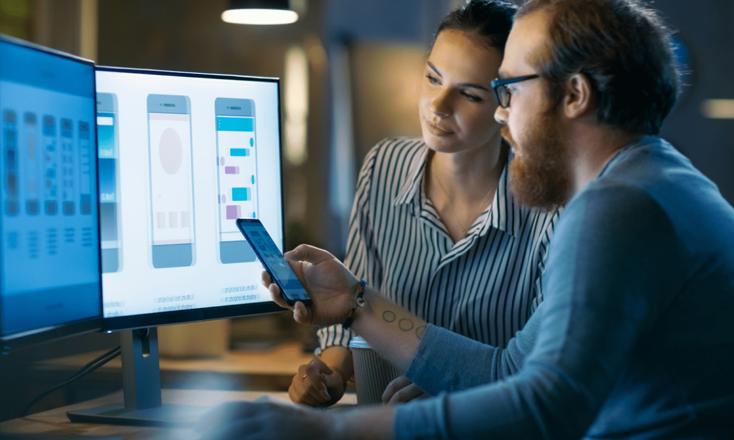 A woman and man in an office comparing an app on a mobile device to several design comps on desktop monitors.