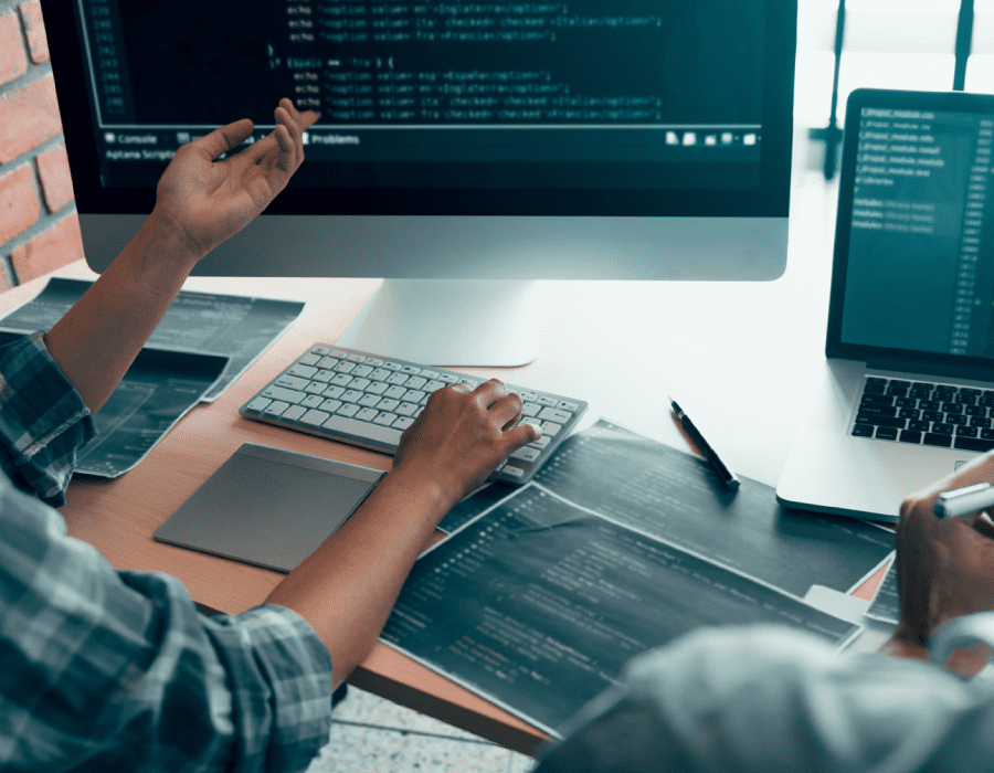 Over the shoulder view of two people sitting at a desk reviewing code on multiple monitors.