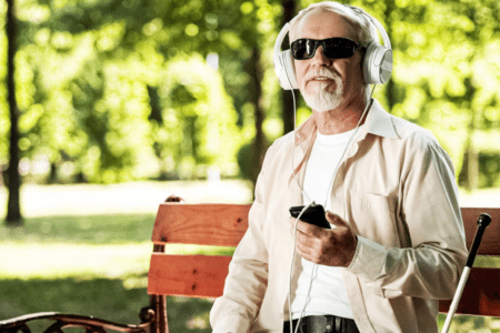 Older man who is blind wearing headphones connected to his mobile device using assistive technology. A white cane is next to him.