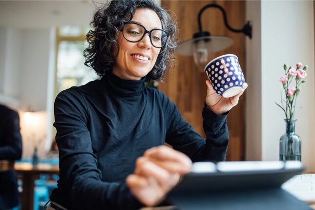 Woman with glasses sitting at a desk working on a laptop while holding a coffee cup.