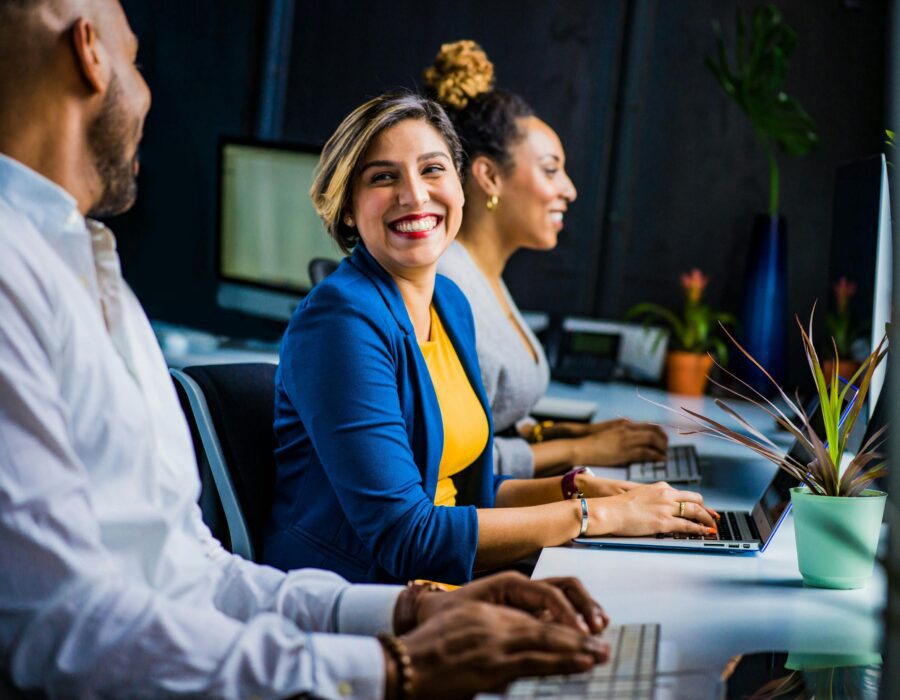 Smiling woman in a blue suit jacket and yellow top working at a laptop next to coworkers