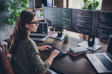 A woman software developer reviews code. Multiple screens sit on her desk.