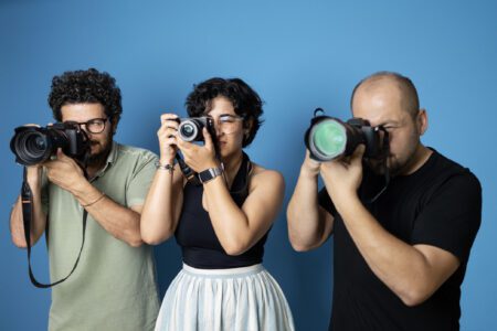 Three photographers, two men and one woman, aiming their cameras while standing in a row against a solid blue background.