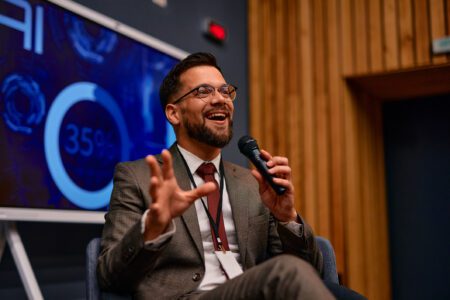 Young businessman sitting and speaking on stage at a business conference, holding a microphone and gesturing with his hands while giving a presentation in front of a screen showing data and charts