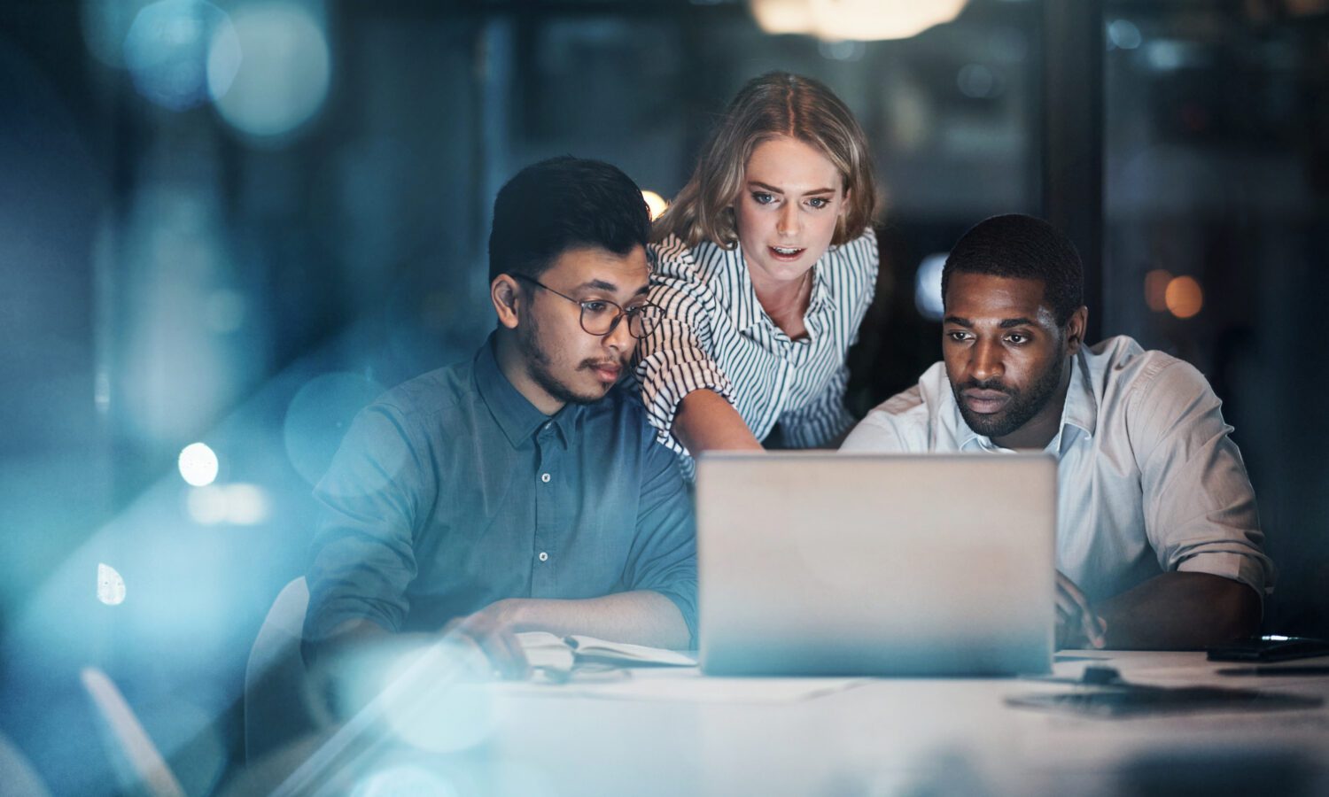 Three colleagues gather around a computer to plan their AI enablement. The room is lit with blue light.