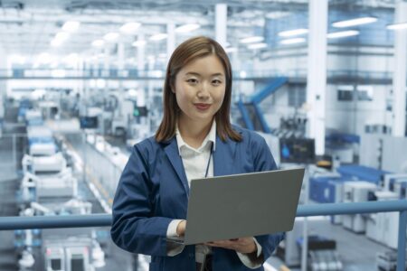 A young female AI software engineer stands with her laptop on a balcony above a factory/warehouse setting.