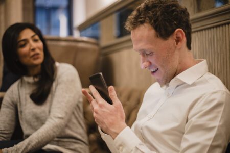 A blind man with his smartphone in his hand is using a screen reader or a mobile app. A woman with dark hair can be seen sitting down next to him.