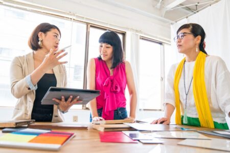 Three women executives stand around a desk or table talking. One holds a tablet and stylus.