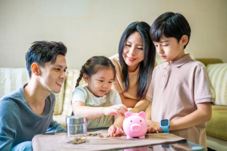 A little girl puts coins into a pink piggy bank while sitting with her family, including an older sibling, in the living room.